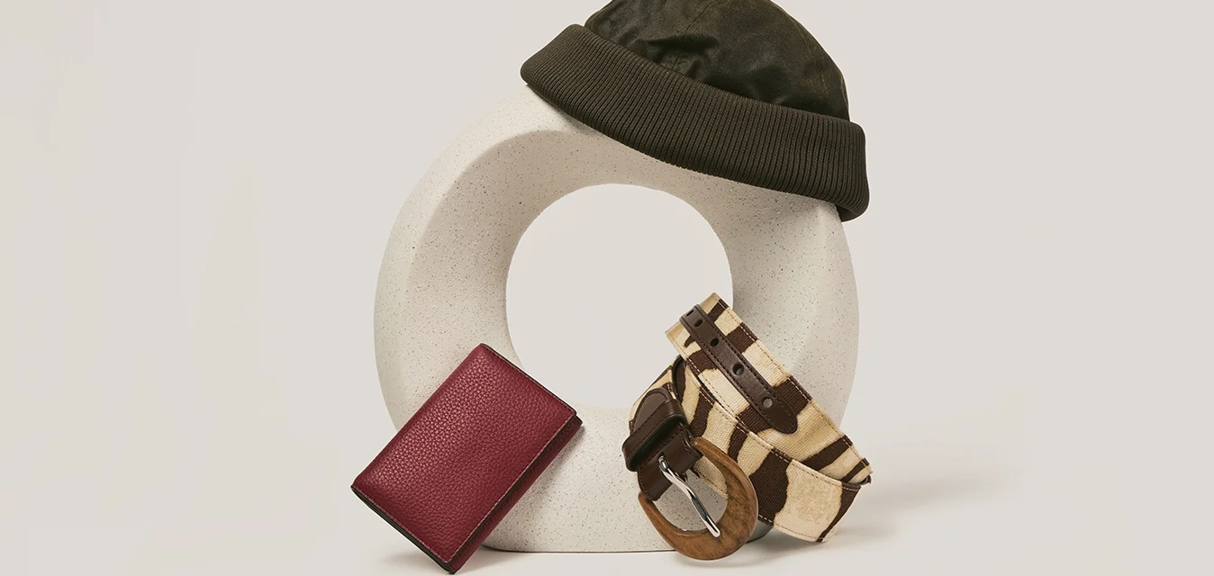 Berretto nero su scultura astratta beige, con un portafoglio rosso e una cintura marrone a righe appoggiati su di essa, su uno sfondo bianco.