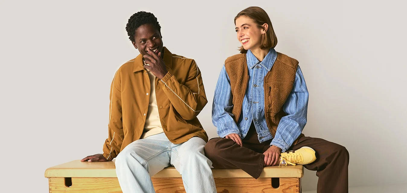 Due persone sedute su una panchina di legno. Una indossa una giacca marrone sopra una camicia beige; l'altra porta una camicia blu e un gilet marrone. Sono visibili delle sneakers.