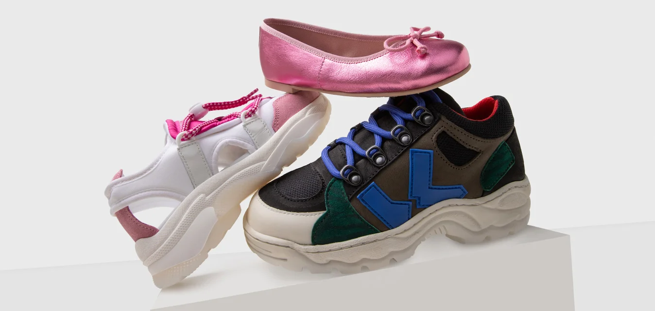 Esposizione di calzature con una ballerina rosa, una scarpa sportiva bianca con dettagli rosa e un sneaker nero con rifiniture blu e verdi.