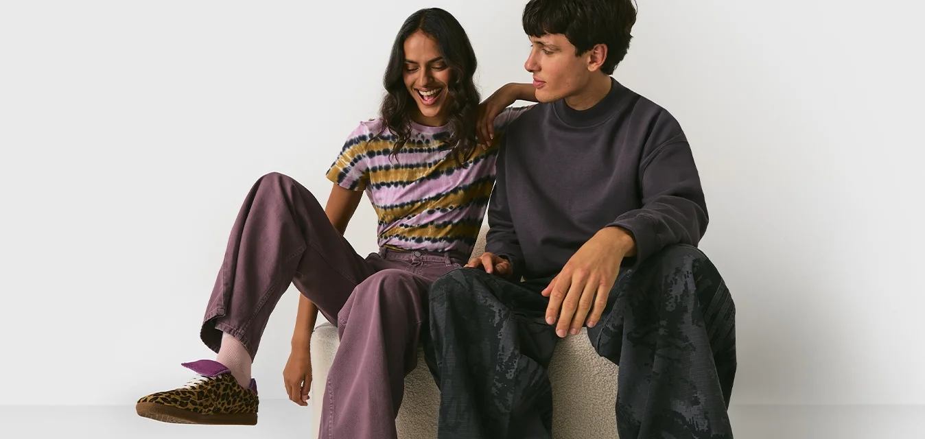 Due giovani adulti in abbigliamento casual seduti su un pouf chiaro, sorridendo, su uno sfondo bianco semplice.