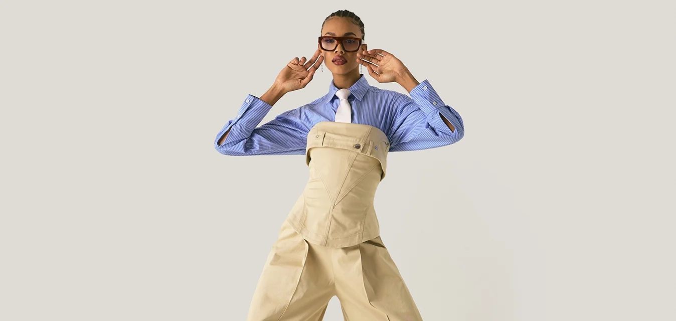 Modella in posa con pantaloni beige a gamba larga e corsetto sopra una camicia a righe blu con cravatta bianca, accessoriata con occhiali e orecchini.
