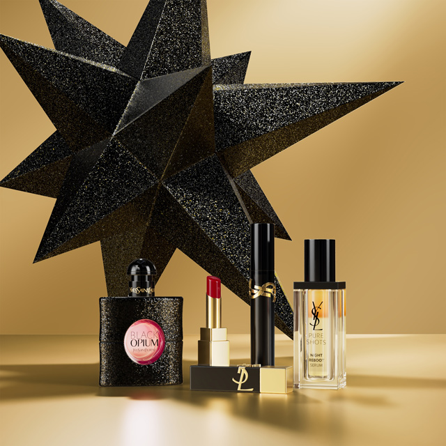 Una composizione di prodotti di bellezza Yves Saint Laurent, inclusi il profumo Black Opium, un rossetto rosso, un mascara e il siero Pure Shots, sono esposti davanti a una grande stella geometrica nera scintillante su una superficie e sfondo dorati.