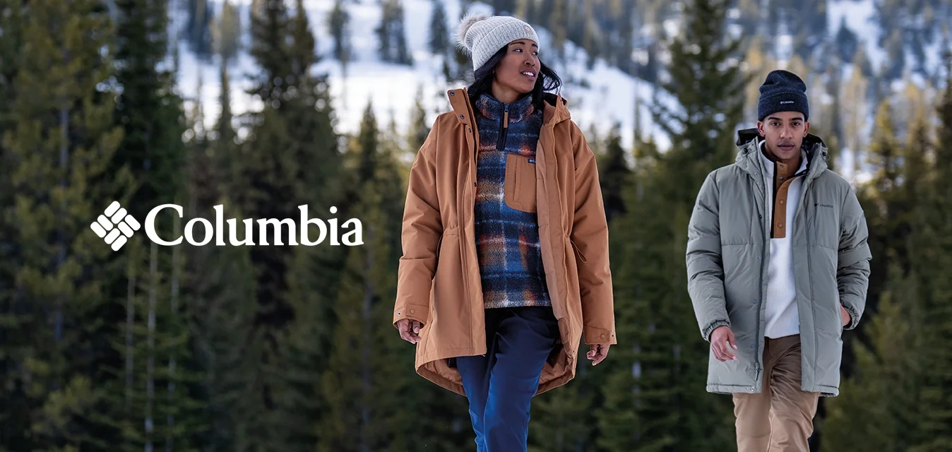 Due persone camminano nella neve, indossando un parka marrone, pantaloni blu, una giacca a vento verde e pantaloni beige. Sullo sfondo si vedono alberi di pino e montagne.