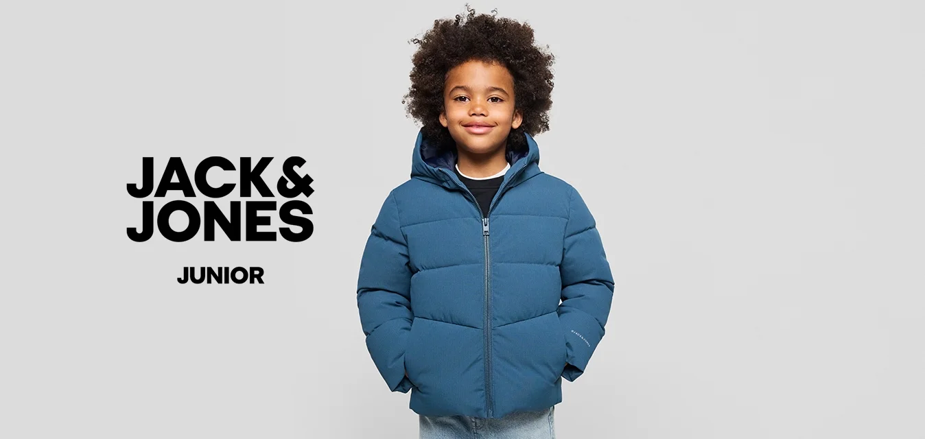 Un bambino con capelli ricci che indossa un giubbotto blu e jeans chiari, in piedi con le mani nelle tasche su uno sfondo neutro.