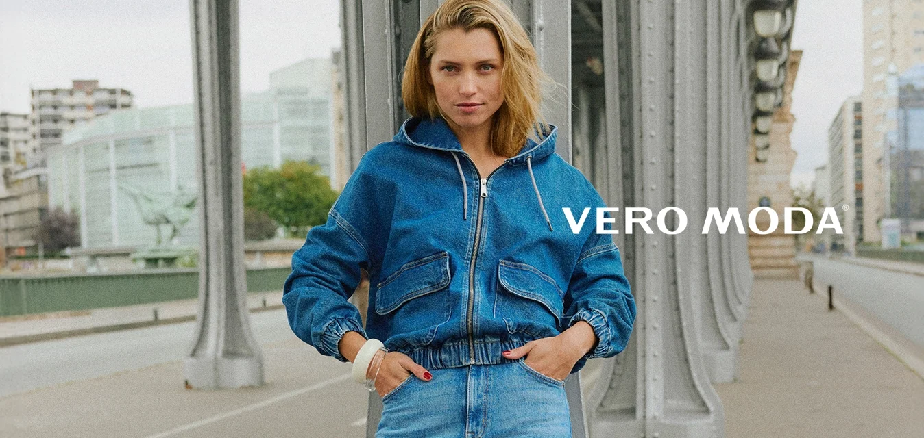 Giovane donna in giacca di denim blu e jeans appoggiata a un pilastro di metallo di un ponte su una strada urbana con edifici moderni sullo sfondo.