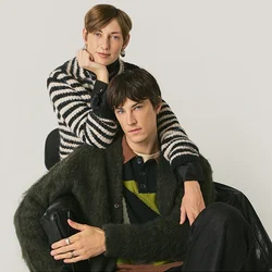 Due giovani adulti, uno seduto e l'altro in piedi dietro con un braccio sulla spalla, entrambi indossano maglioni a trama e a righe su uno sfondo semplice.