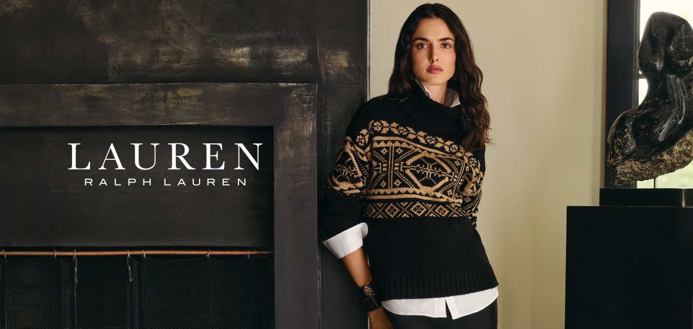 Donna in nero, maglione con motivo beige e lunga gonna nera si trova vicino a un caminetto scuro e a una scultura astratta su un piedistallo vicino alla finestra.