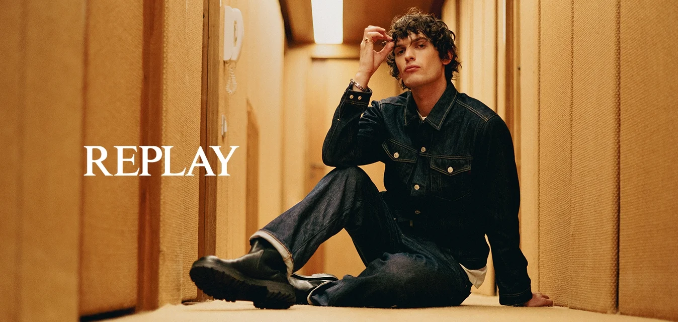 Giovane in giacca di jeans e pantaloni di denim è seduto sul pavimento di moquette di un stretto corridoio beige, toccandosi i capelli con una mano.