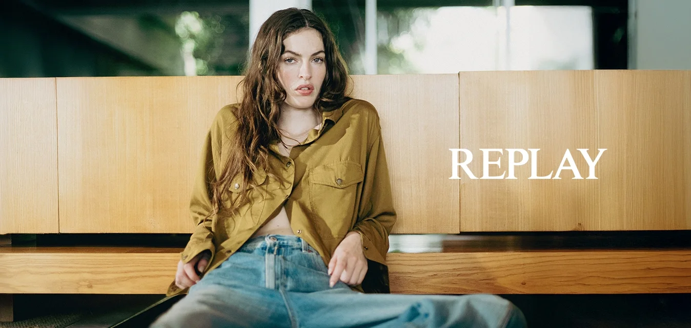 Giovane donna con una camicia verde oliva e jeans blu, seduta per terra appoggiata a un mobile di legno all'interno.