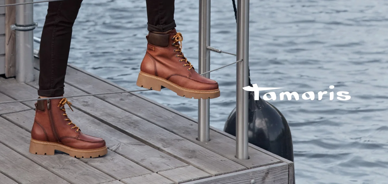 Cappotto impermeabile marrone con colletto, maglione crema, pantaloni neri e stivaletti marroni con lacci e suola spessa. Contesto in legno e metallo.