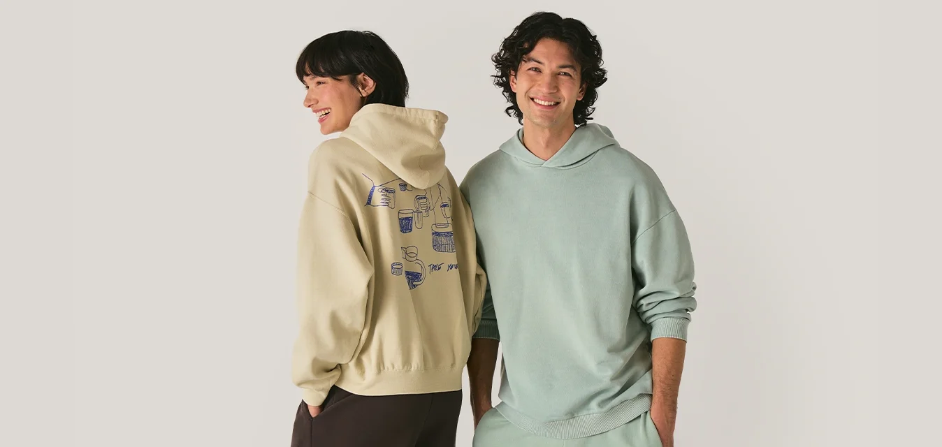 Uomo e donna sorridenti che indossano felpe pastello e pantaloni comodi, in piedi fianco a fianco contro uno sfondo chiaro e semplice.