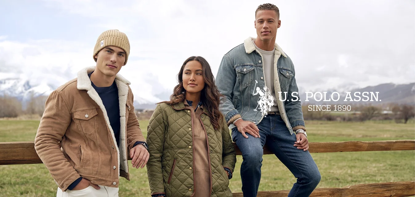 Tre giovani adulti vestiti con giacche invernali e stivali posano accanto a una recinzione di legno in un campo erboso, con montagne innevate sullo sfondo.