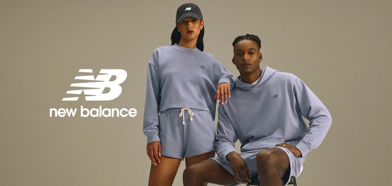 Due modelli in abbigliamento sportivo viola chiaro abbinato; uno in piedi con un cappello, l'altro seduto su uno sgabello nero, entrambi con scarpe bianche.