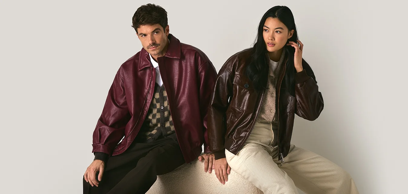 Un uomo e una donna seduti su una panchina curva, indossano giacche di pelle, l'uomo in bordeaux e la donna in marrone scuro, con sfondo neutro.