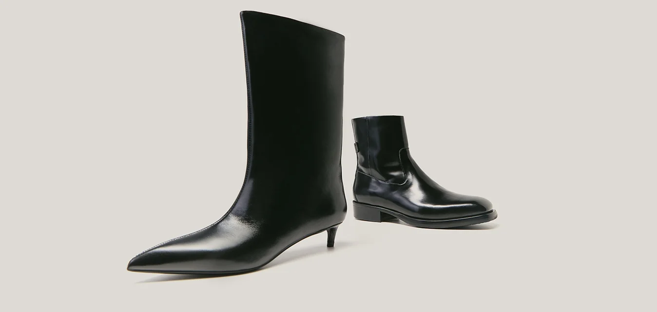 Due stivali neri in pelle isolati su sfondo chiaro; uno alto con punta a punta e tacco kitten, l'altro un stivaletto corto con suola piatta.