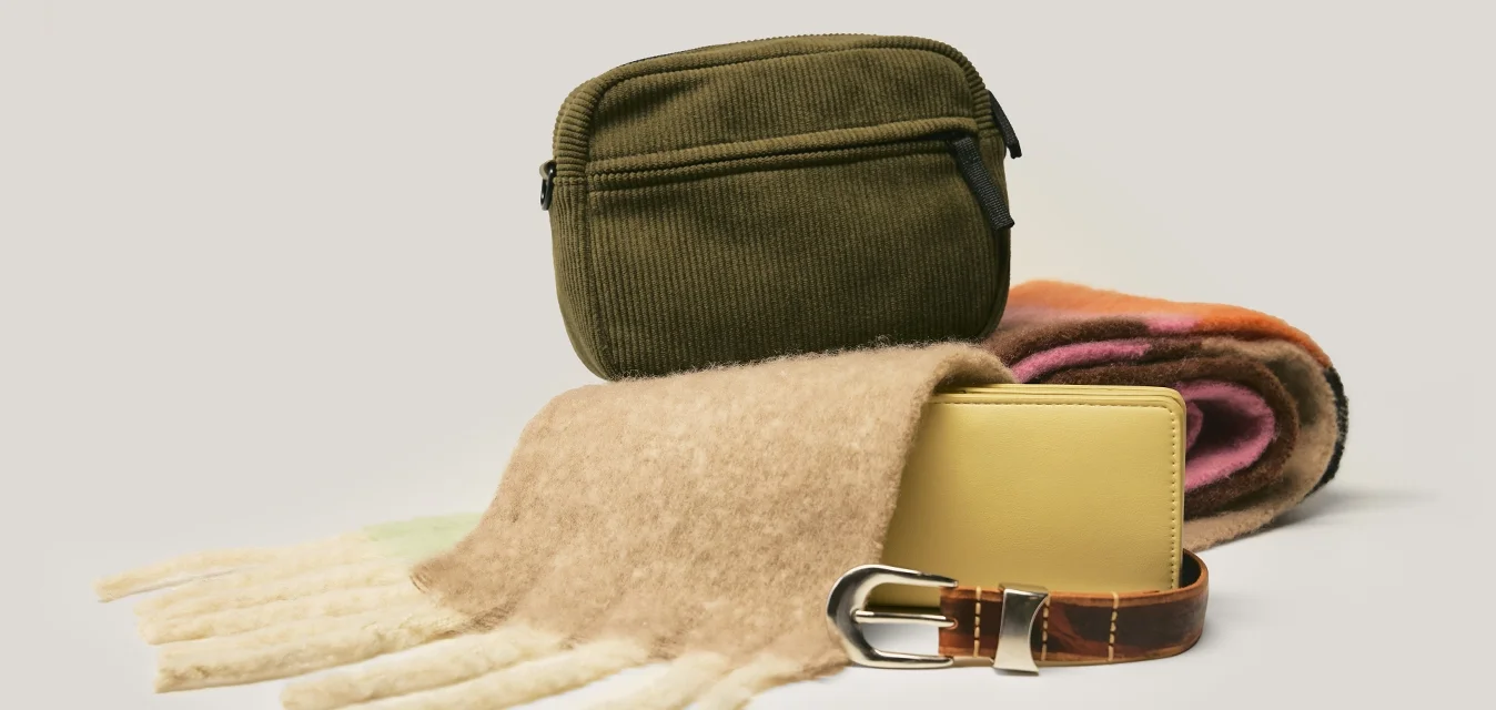 Borsa in velluto a coste verde, sciarpa beige morbida, portafoglio giallo e cintura in pelle marrone con fibbia metallica su uno sfondo neutro.
