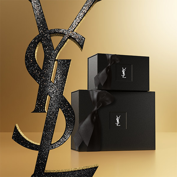 Due eleganti scatole regalo nere YSL con nastri sono appoggiate su uno sfondo dorato con un grande logo YSL nero scintillante.