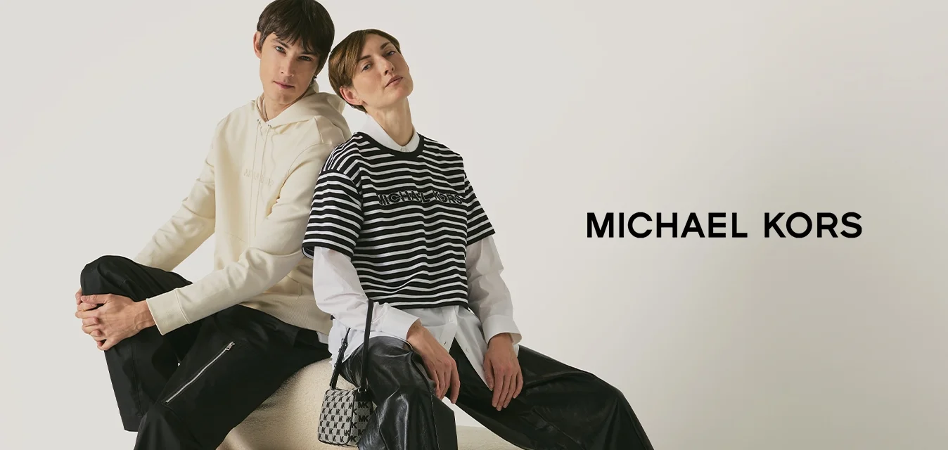 Due giovani seduti schiena contro schiena su un ottomano rotondo beige, vestiti con outfit casual chic, su uno sfondo bianco.