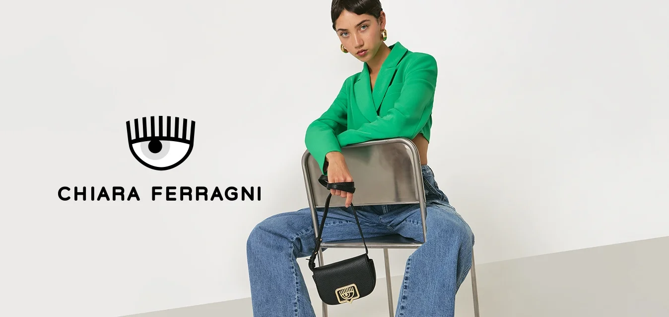 Donna seduta all'indietro su una sedia di metallo, con un blazer verde, jeans larghi blu, tacchi bianchi e sandali con cinturino, mentre tiene una piccola borsa nera.