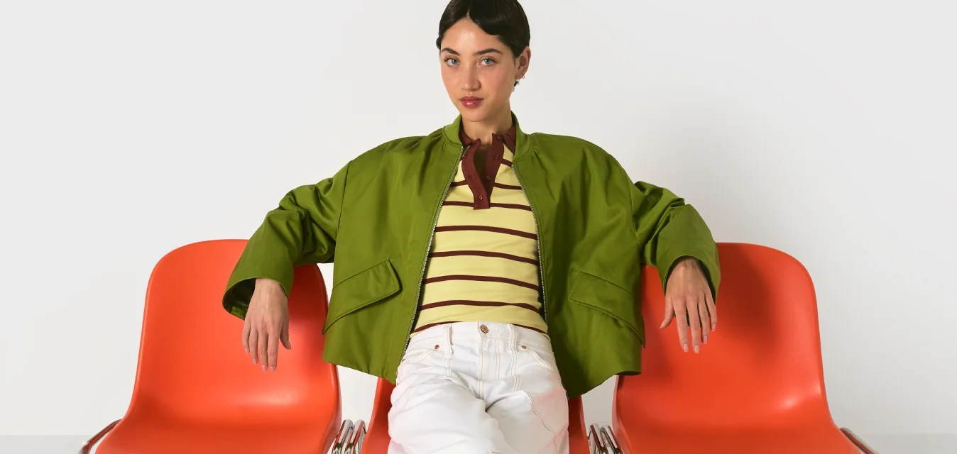 Donna seduta con le gambe incrociate su una sedia arancione a metà, con le braccia appoggiate su sedie vuote adiacenti, indossa una giacca verde e scarpe rosse.