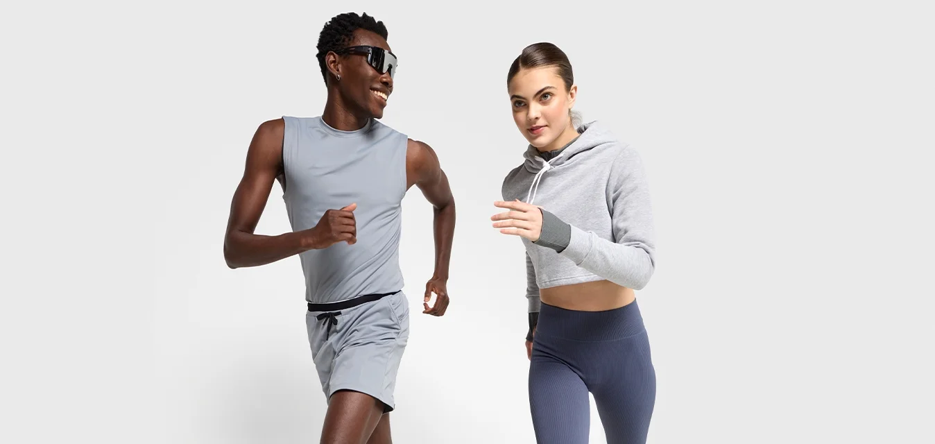 Due persone che corrono indoors indossando abbigliamento sportivo; una in pantaloni corti grigi e top senza maniche, l'altra in leggings blu navy e felpa grigia corta.