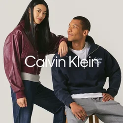 Giovane donna con giacca bordeaux e jeans sta accanto a un uomo in felpa blu navy e pantaloni grigi seduto su uno sgabello contro uno sfondo semplice.