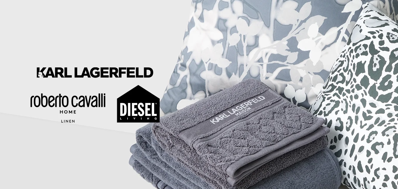 Pila di asciugamani grigi Karl Lagerfeld su una superficie bianca con cuscini a stampa floreale e leopardata sullo sfondo.