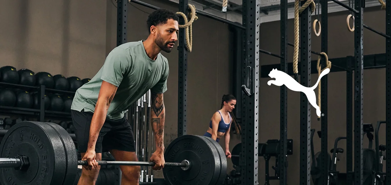 Cinque persone che si allenano in una palestra; uno solleva un bilanciere, gli altri utilizzano palle mediche o attrezzature da palestra in un'area di allenamento ampia e ben illuminata.