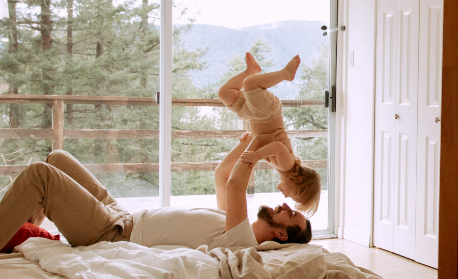 alt="Padre sdraiato su un letto che solleva scherzosamente il suo bambino in una camera da letto illuminata dal sole con vista sulla foresta fuori dalla finestra.»