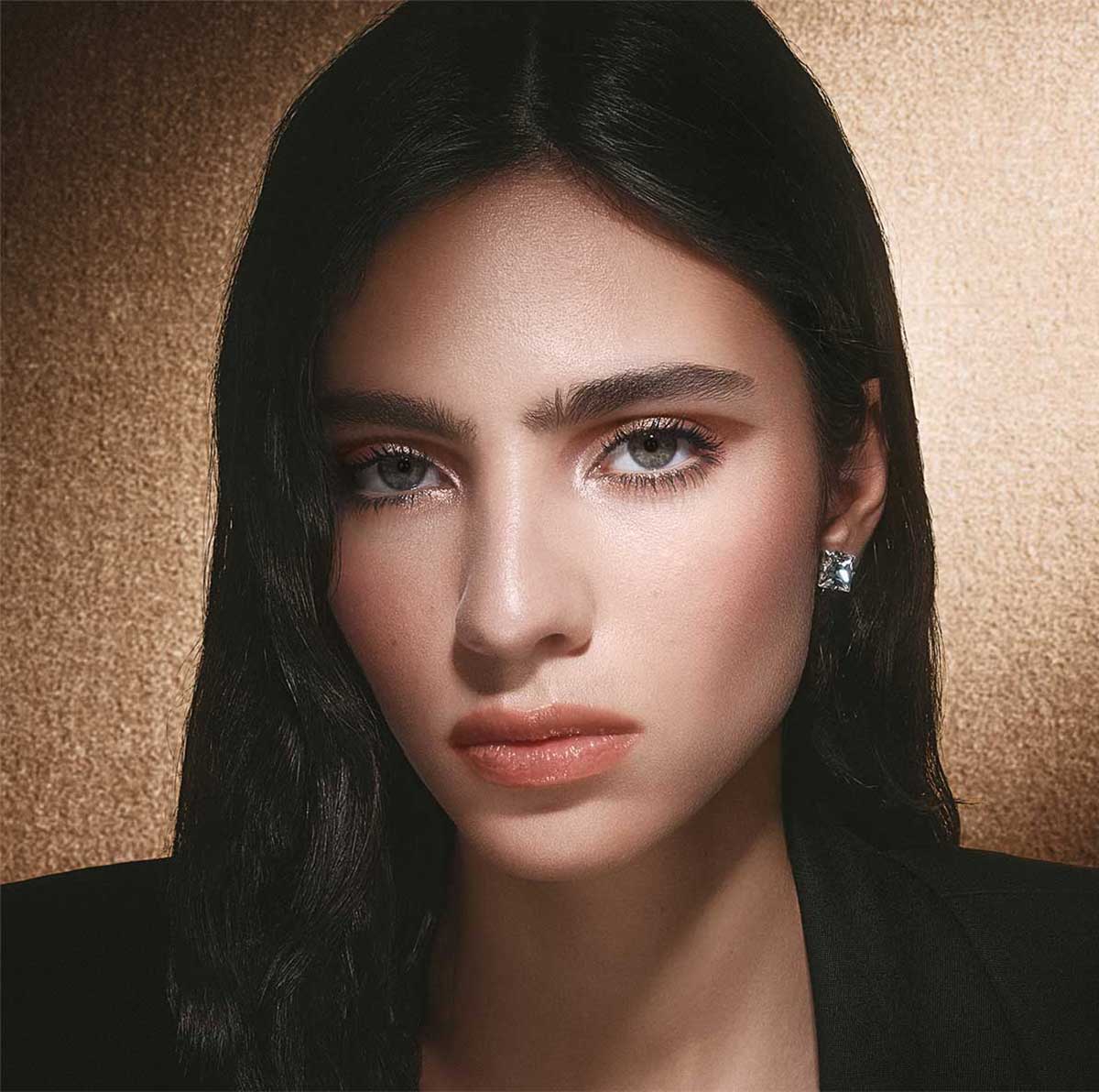 Un primo piano di una donna con lunghi capelli scuri, che indossa un blazer nero, un trucco occhi discreto e un colore labbra naturale, su uno sfondo dorato scintillante.