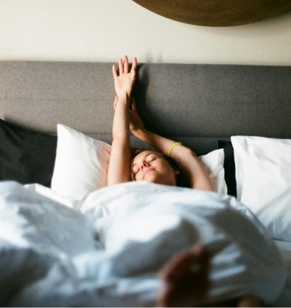 alt="Donna che si sveglia e allunga le braccia in un letto luminoso e accogliente con lenzuola bianche.»