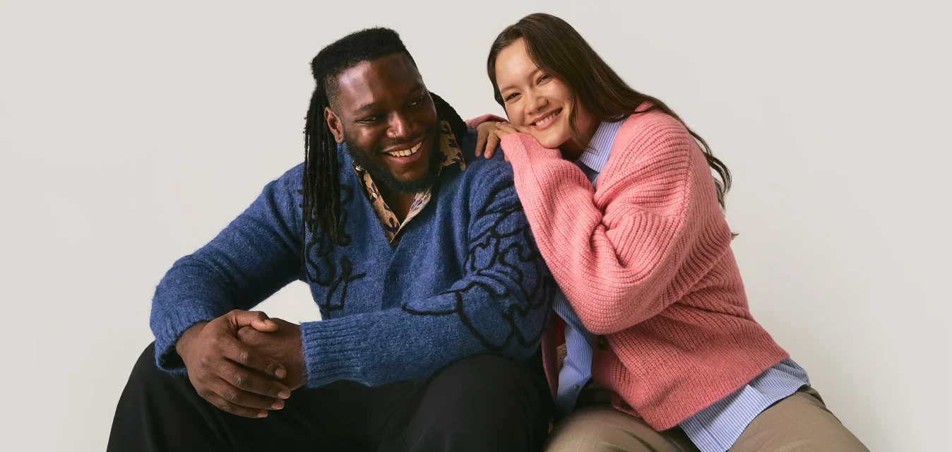 Uomo sorridente in maglione blu e donna in maglione rosa siedono vicini, la donna si appoggia sulla spalla dell'uomo su uno sfondo chiaro e semplice.
