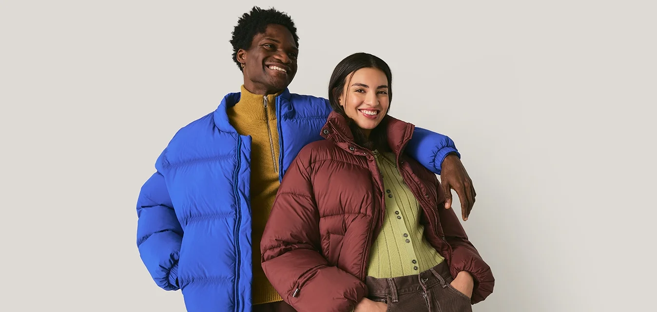 Due adulti sorridenti con giacche invernali colorate, in piedi vicini con le mani in tasca davanti a uno sfondo neutro.