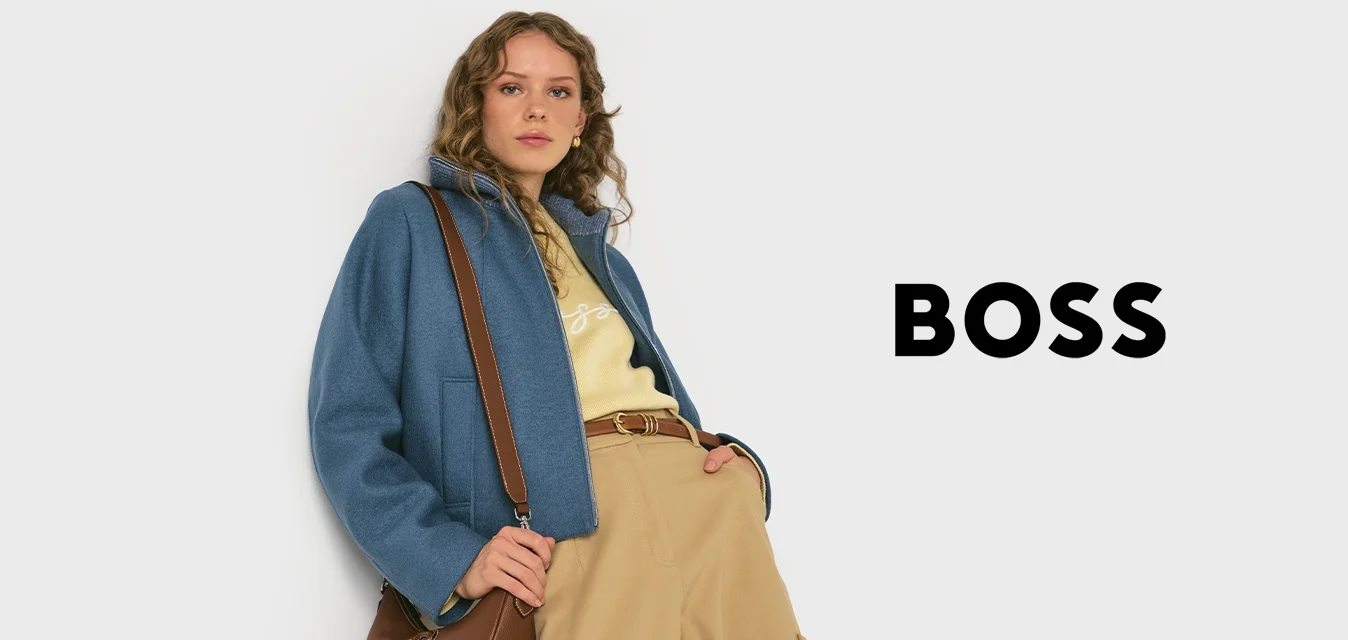 Donna che indossa una giacca blu, pantaloni beige e un top giallo, mentre tiene una borsa a tracolla marrone, in piedi davanti a uno sfondo semplice.