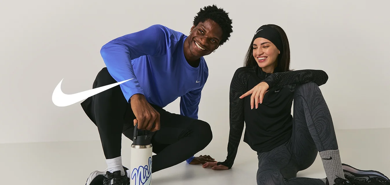 Due persone in abbigliamento sportivo sedute a terra, sorridenti; una tiene una bottiglia d'acqua, entrambe sembrano rilassate e amichevoli.
