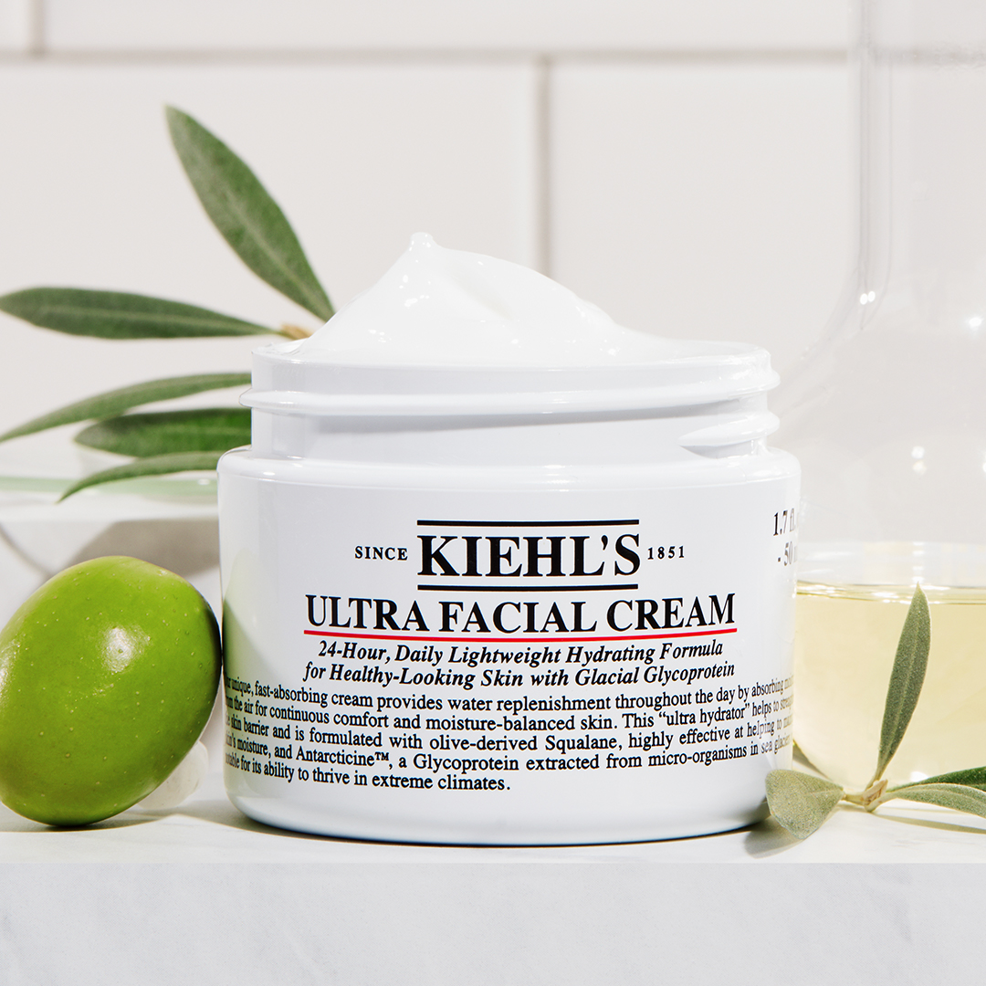 Vaso aperto di Ultra Facial Cream su sfondo luminoso con ingredienti botanici disposti intorno.