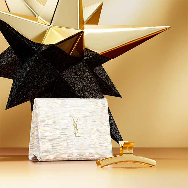 Una pochette YSL Beauté bianca e oro e un fermaglio per capelli dorato sono esposti contro uno sfondo di grandi decorazioni geometriche a forma di stella dorate e nere scintillanti.