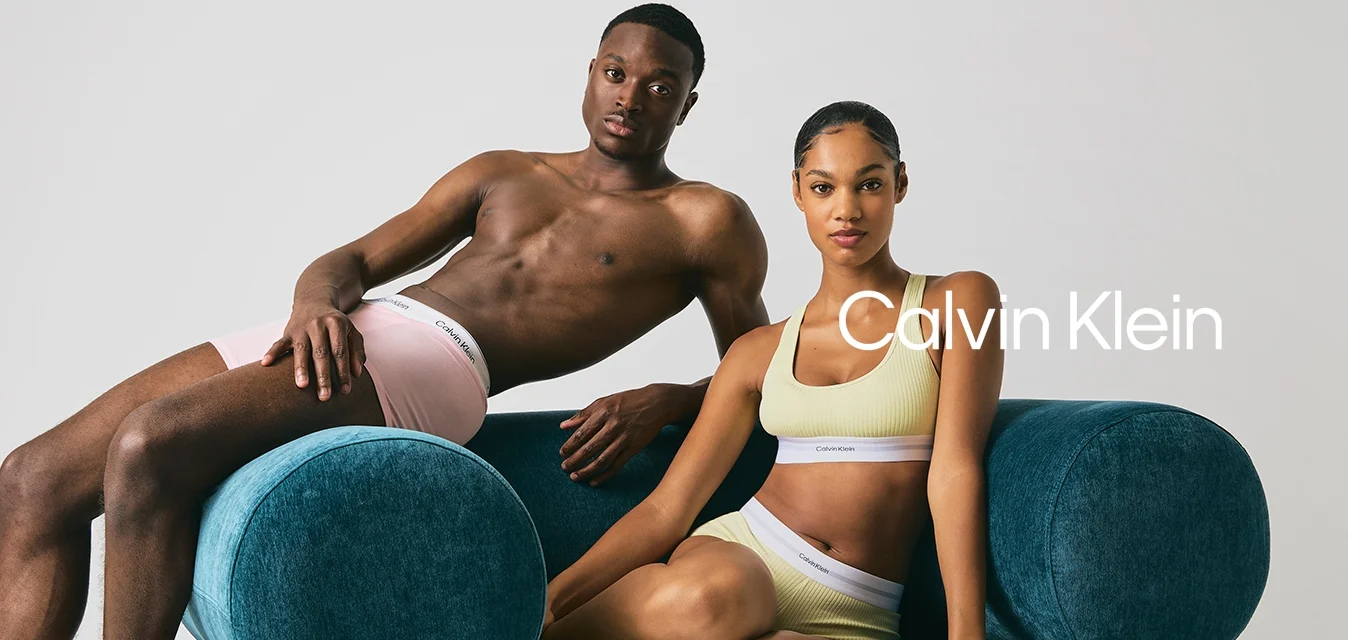 Uomo in boxer rosa che si sdraia sul bracciolo di un divano blu arrotondato, donna in reggiseno sportivo giallo a coste e pantaloni corti che si siede accanto a lui.