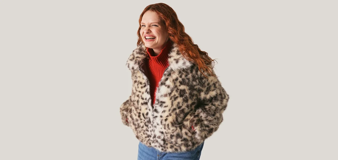 Giovane donna con capelli rossi sorride, indossa una giacca di pelliccia con stampa leopardata, un maglione dolcevita rosso e jeans blu, con le mani nelle tasche.