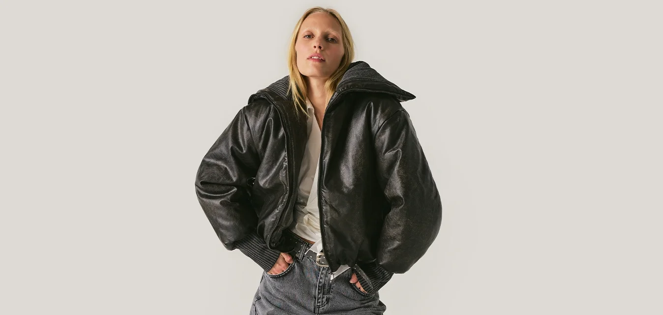 Donna che indossa una giacca nera oversize lucida, camicia bianca e pantaloni a righe a gamba larga, in piedi con le mani nelle tasche davanti a uno sfondo neutro.