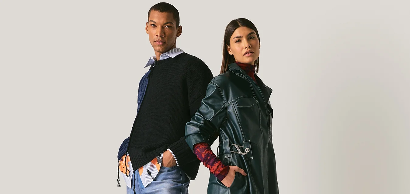 Uomo e donna in piedi schiena contro schiena, vestiti con abiti moderni a strati con denim, trench in pelle e scarpe casual su sfondo neutro.