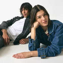 Due persone sono sdraiate su una superficie bianca. Una indossa una giacca di jeans nera sopra una camicia bianca, mentre l'altra indossa una giacca di jeans blu sopra una maglietta a righe.