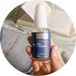 alt="Con in mano una bottiglia di spray liposomiale GABA + L-Teanina Quicksilver Scientific, con un libro aperto e biancheria da letto accogliente sullo sfondo.»