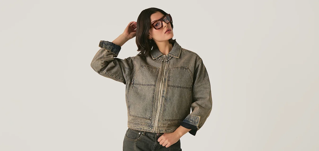 Donna che indossa un giubbotto di jeans oversize e una gonna abbinata, mentre sistema i capelli su uno sfondo neutro.