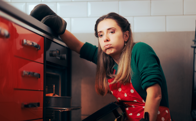 alt="Donna in grembiule seduta stancamente davanti a un forno, appoggia il braccio sul bancone, esprimendo stress e affaticamento.»