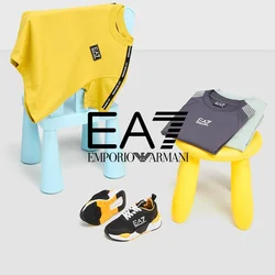 Due magliette per bambini, una gialla e una grigia, esposte su sgabelli colorati. Sneakers nere e arancioni sono posizionate nei pressi sul pavimento.