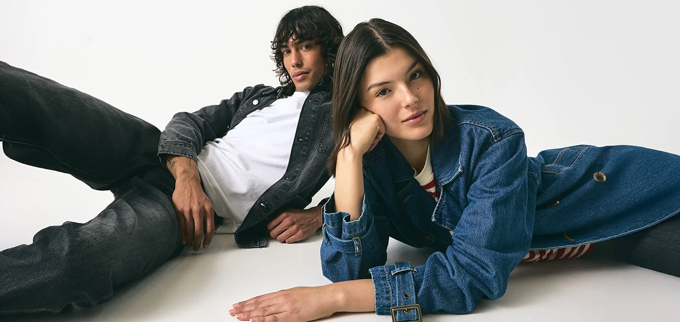 Due persone sono sdraiate su una superficie bianca. Una indossa una giacca di jeans nera sopra una camicia bianca, mentre l'altra indossa una giacca di jeans blu sopra una maglietta a righe.