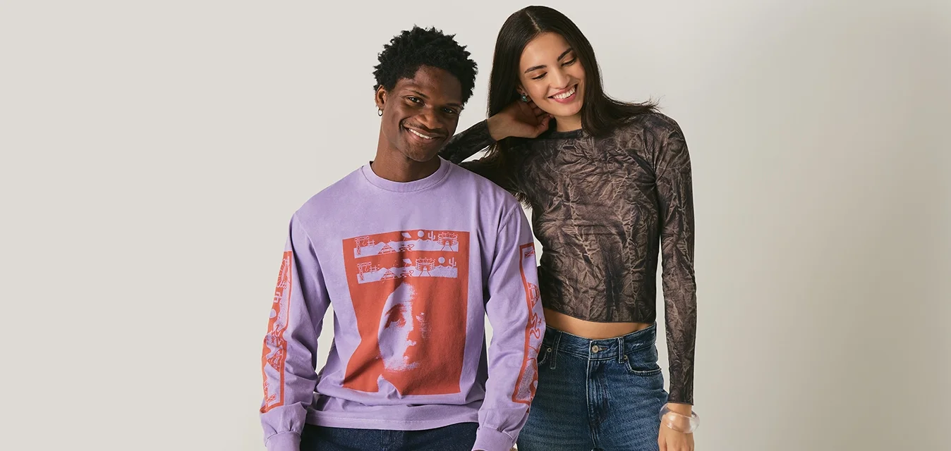 Uomo con maglione viola a motivo grafico seduto su uno sgabello accanto a una donna sorridente con maglia a maniche lunghe a motivi e jeans blu, su sfondo semplice.