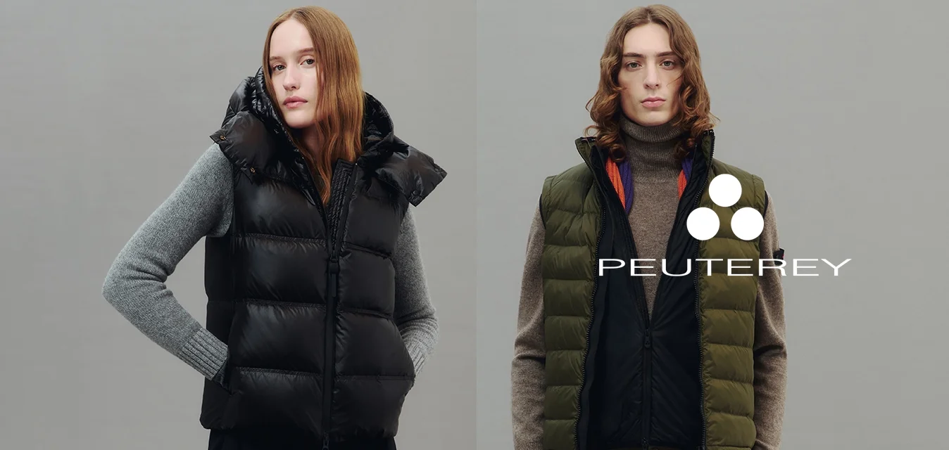 Due modelli indossano gilet senza maniche imbottiti sopra maglioni a maniche lunghe, uno di colore nero con cappuccio, l'altro di colore verde oliva con dolcevita marrone.