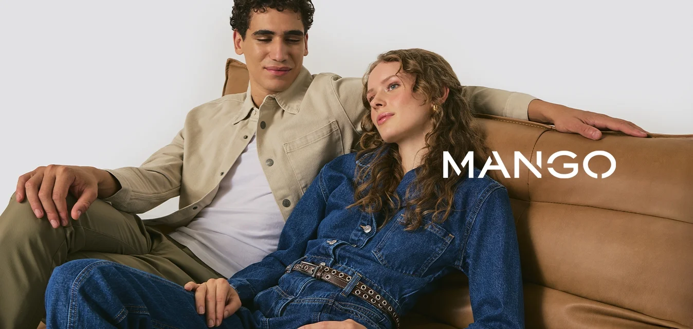 Due persone sono sedute su un divano in pelle marrone chiaro. Una indossa una giacca beige sopra una camicia bianca, l'altra un tuta di jeans blu con una cintura.